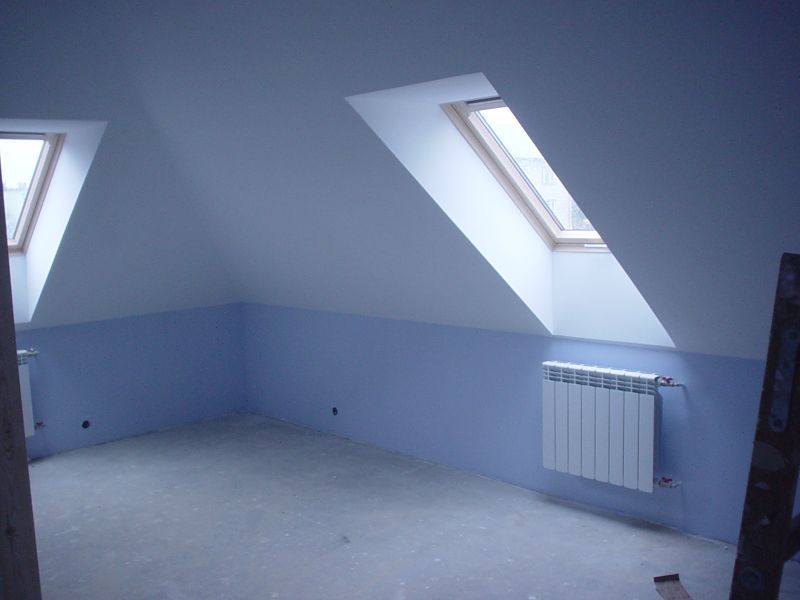 Pokój na poddaszu po remoncie, z dwoma oknami dachowymi, pomalowany na biało i błękitno, z grzejnikiem na ścianie. Widoczne ślady po gniazdkach elektrycznych. Podłoga surowa, betonowa.