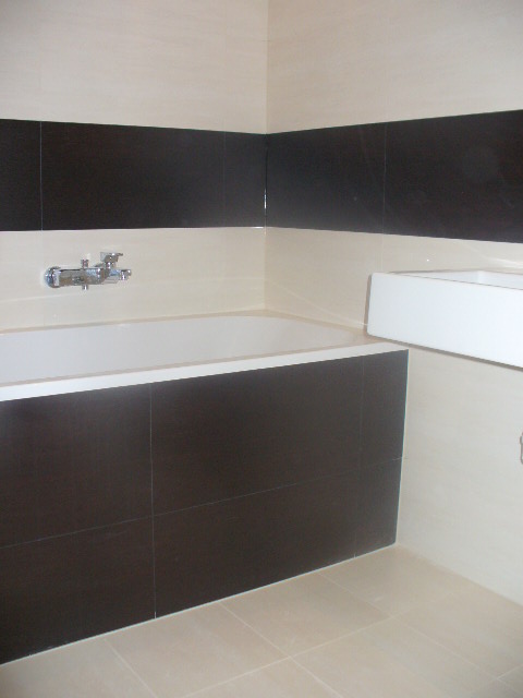 Wykończona łazienka z wanną obudowaną ciemnymi płytkami, jasne płytki na ścianach i podłodze, bateria wannowa chromowana.