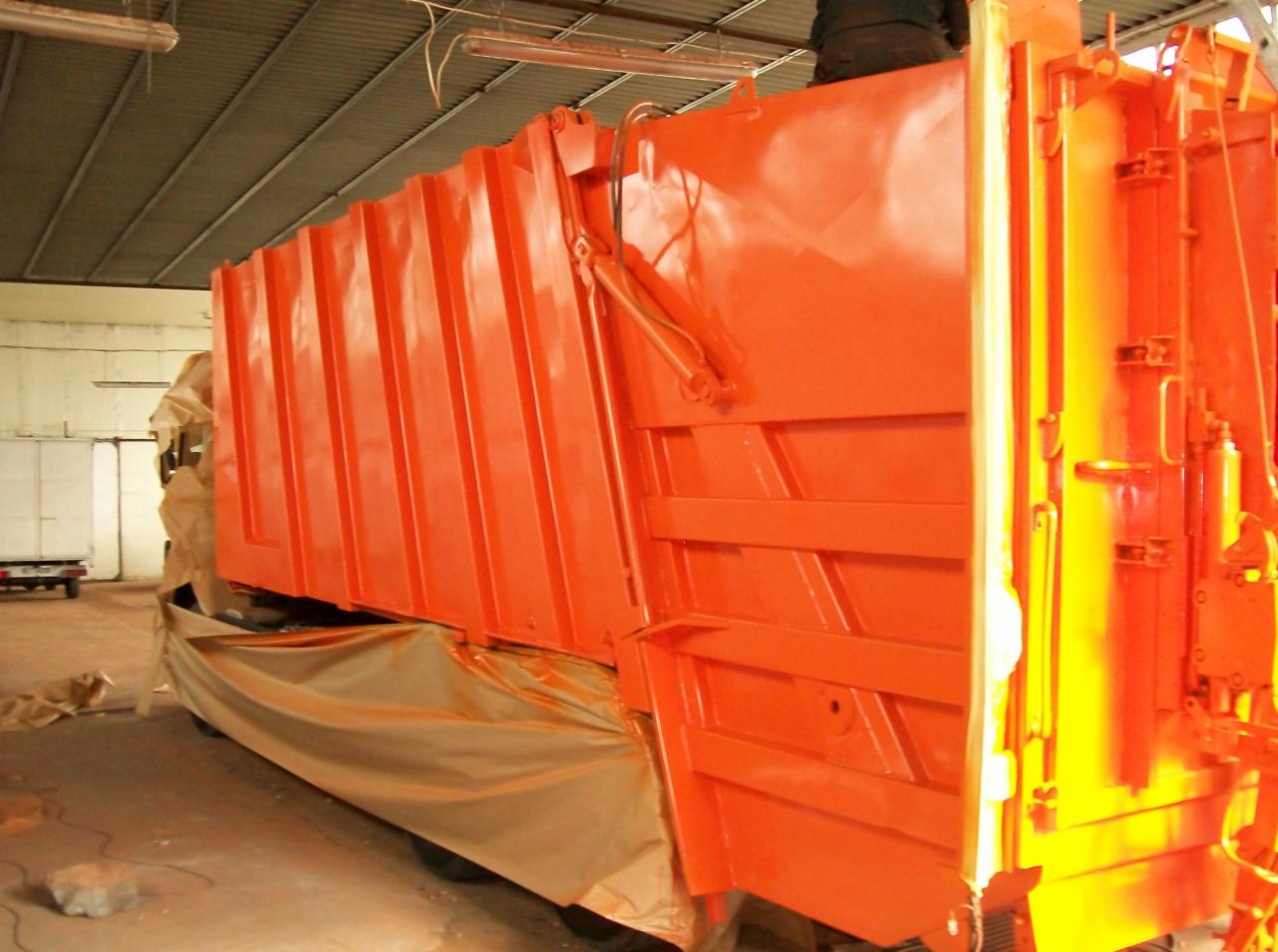 Pomarańczowy kontener śmieciarki po malowaniu w hali, z częściowo zamaskowanym podwoziem i widocznymi elementami hydraulicznymi.