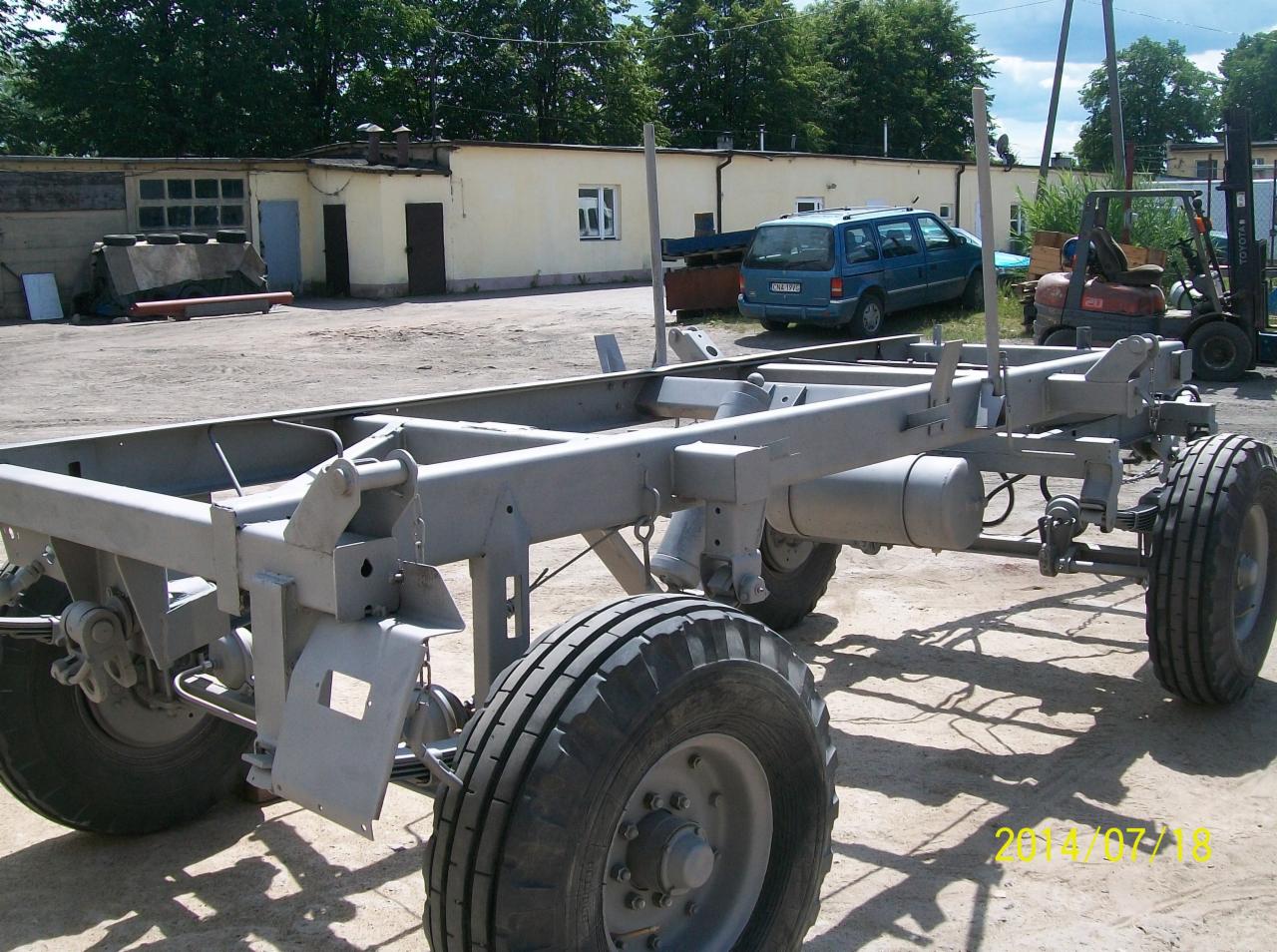 Malowana na szaro rama pojazdu z kołami na zewnątrz, z budynkiem, samochodem i wózkiem widłowym w tle, w słoneczny dzień.