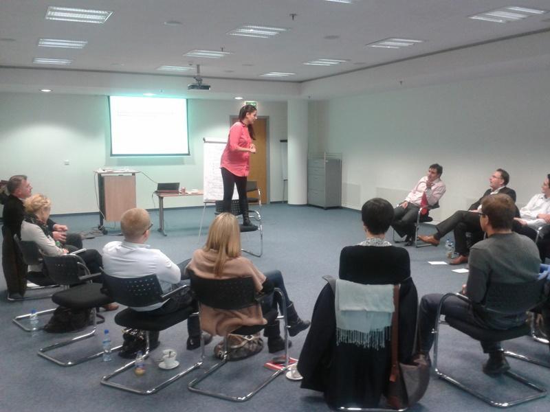 Prelegentka w różowej koszuli stoi na krześle podczas prezentacji dla grupy osób siedzących na krzesłach w sali konferencyjnej z projektorem i ekranem.