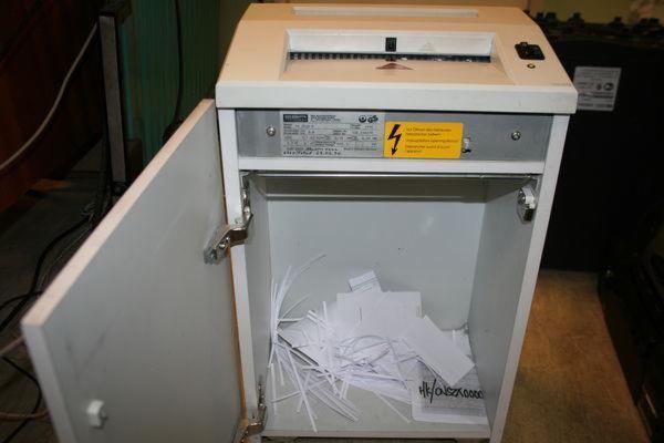 Biała niszczarka biurowa z otwartymi drzwiczkami, wypełniona pociętymi dokumentami. Widoczna żółta naklejka ostrzegawcza i metalowa tabliczka znamionowa.