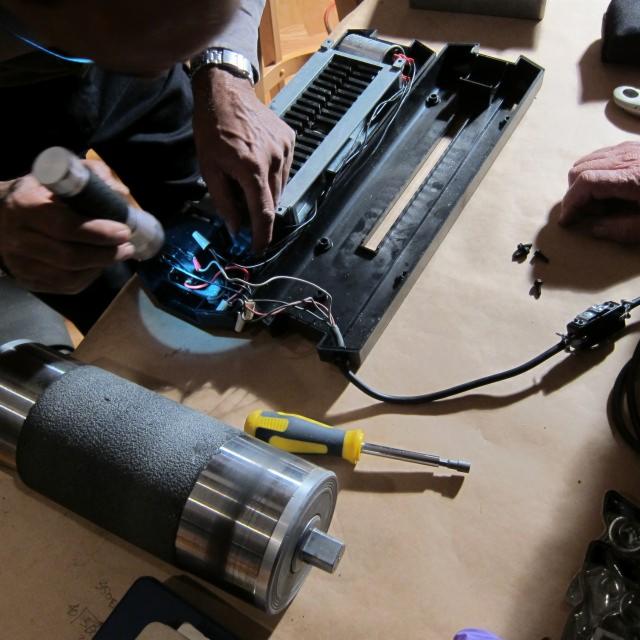 Serwisant naprawia rozłożony sprzęt biurowy oświetlając wnętrze latarką. Widoczne narzędzia: śrubokręt i elementy elektroniczne.