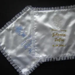 Dwie białe poduszki z satyny z haftem imion i dat chrztu, wykończone falbanką, leżące na ciemnym tle.