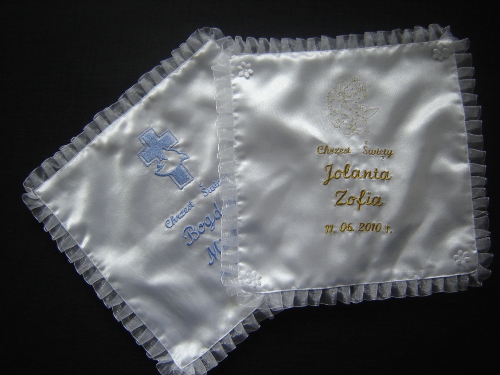 Dwie białe poduszki z satyny z haftem imion i dat chrztu, wykończone falbanką, leżące na ciemnym tle.