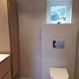 Nowoczesna, minimalistyczna łazienka z podwieszaną toaletą, jasnymi płytkami ściennymi i szafką z drewna w ciepłym odcieniu, małe okno z widokiem na zieleń.