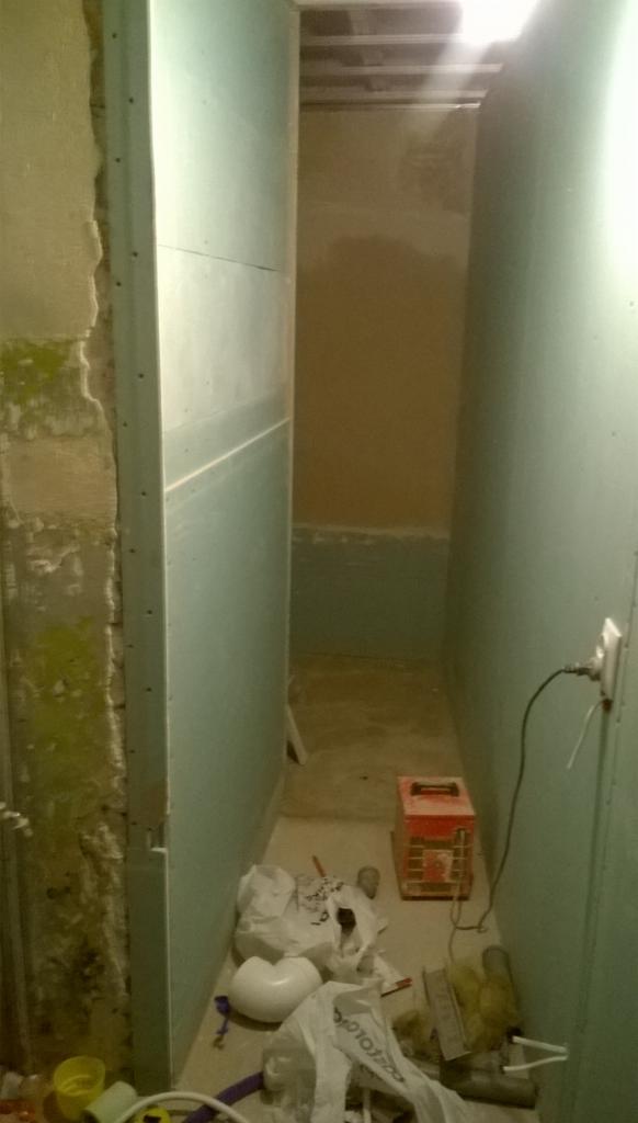 Wąskie przejście w trakcie remontu: ściana z surowego betonu z zielonymi plamami, obok płyta karton-gips na metalowym profilu, na podłodze gruz, śmieci i narzędzia.