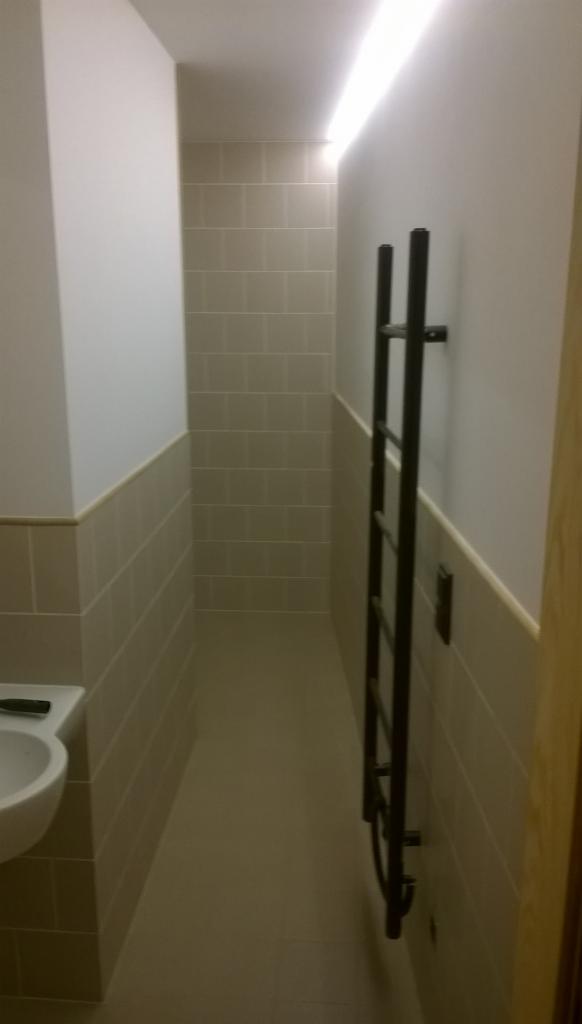 Wąska łazienka po remoncie z beżowymi płytkami do połowy ściany, grzejnikiem drabinkowym i podświetleniem sufitowym LED.