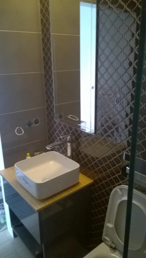 Nowoczesna łazienka z umywalką nablatową na drewnianym blacie, grafitowe płytki w marokański wzór na ścianie, lustro z półką i biała toaleta w tle.