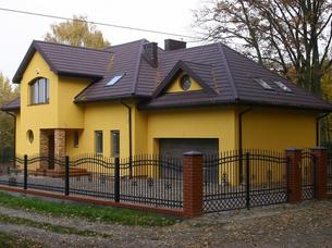Żółty dom jednorodzinny z brązowym dachem i czarnym, metalowym ogrodzeniem z ceglanymi słupkami. Widoczne okna o różnych kształtach i komin. Tło stanowią drzewa z jesiennymi liśćmi.