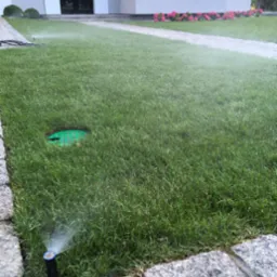 Automatyczny system nawadniania trawnika w trakcie pracy, z widocznymi dyszami rozpylającymi wodę na tle domu z kwiatami.