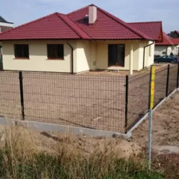 Nowoczesne, czarne panele ogrodzeniowe okalają posesję z beżowym domem i czerwoną dachówką. Widoczne słupki z żółtymi oznaczeniami. W tle zabudowa jednorodzinna.