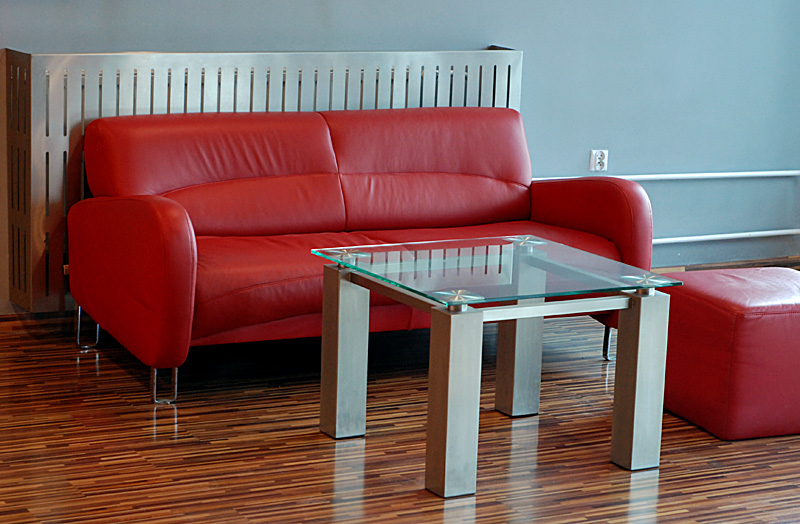 Czerwona skórzana sofa i pufa z kwadratowym stolikiem kawowym ze szklanym blatem i stalową ramą na parkiecie, na tle szarej ściany z osłoną grzejnika ze stali nierdzewnej.