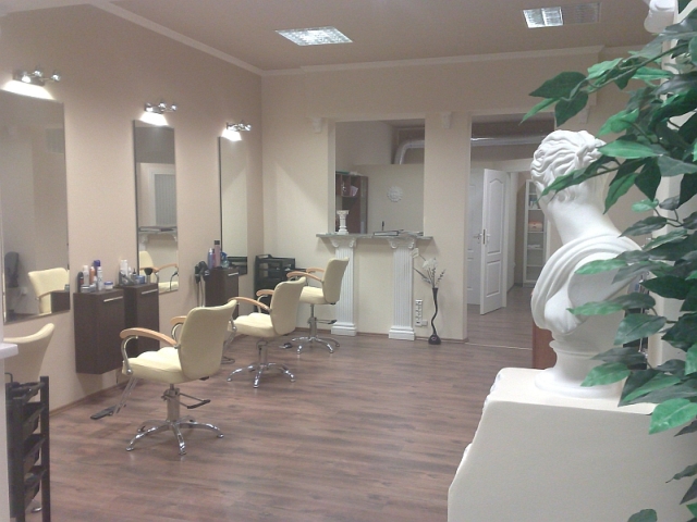 Wnętrze lokalu użytkowego po remoncie: brązowa podłoga, kremowe ściany, stanowiska fryzjerskie z lustrami i oświetleniem, dekoracyjna rzeźba z rośliną w rogu.