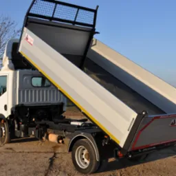 Biała wywrotka z otwartą skrzynią ładunkową, widoczna z tyłu pod kątem, na tle polnego krajobrazu i błękitnego nieba.