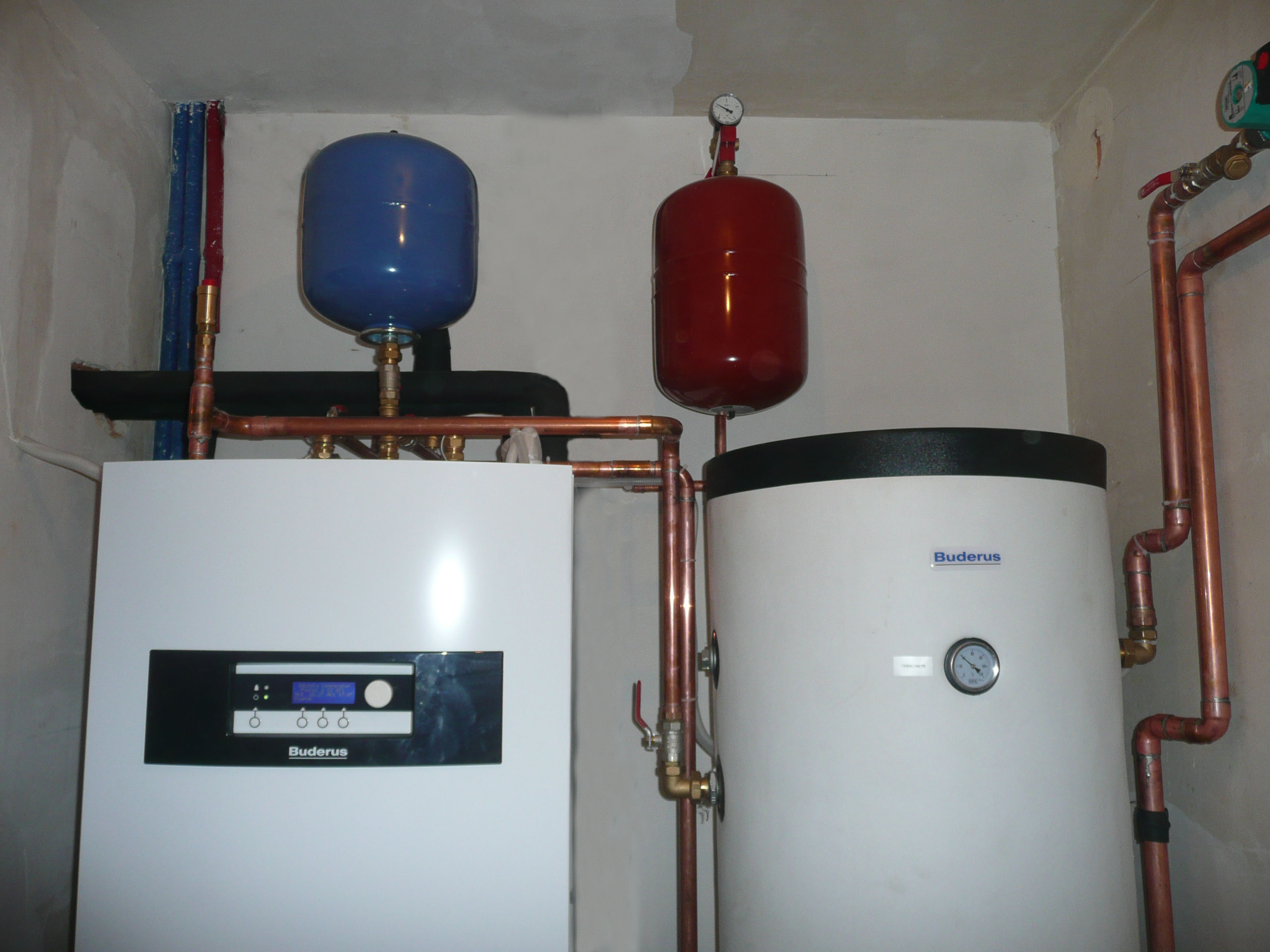 Biały piec grzewczy Buderus z niebieskim i czerwonym naczyniem przeponowym, miedziane rury instalacji CO, widoczne manometry i fragment ściany w surowym stanie.
