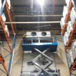 Pracownik na podnośniku nożycowym montuje agregat chłodniczy w magazynie z wysokimi regałami.