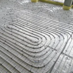 Rozłożone rury ogrzewania podłogowego na folii z siatką, widoczne mocowania rur, fragment okna z ociepleniem w tle.