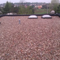 Dach płaski pokryty drobnym żwirem w odcieniach brązu i szarości, z dwoma świetlikami i elementami wentylacyjnymi. W tle widoczne zabudowania mieszkalne.