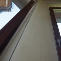 Beżowa ściana z oknami o brązowych ramach, widok z dołu. Delikatna struktura tynku i oświetlenie podkreślają wykończenie wnętrza. Geometryczny układ okien.