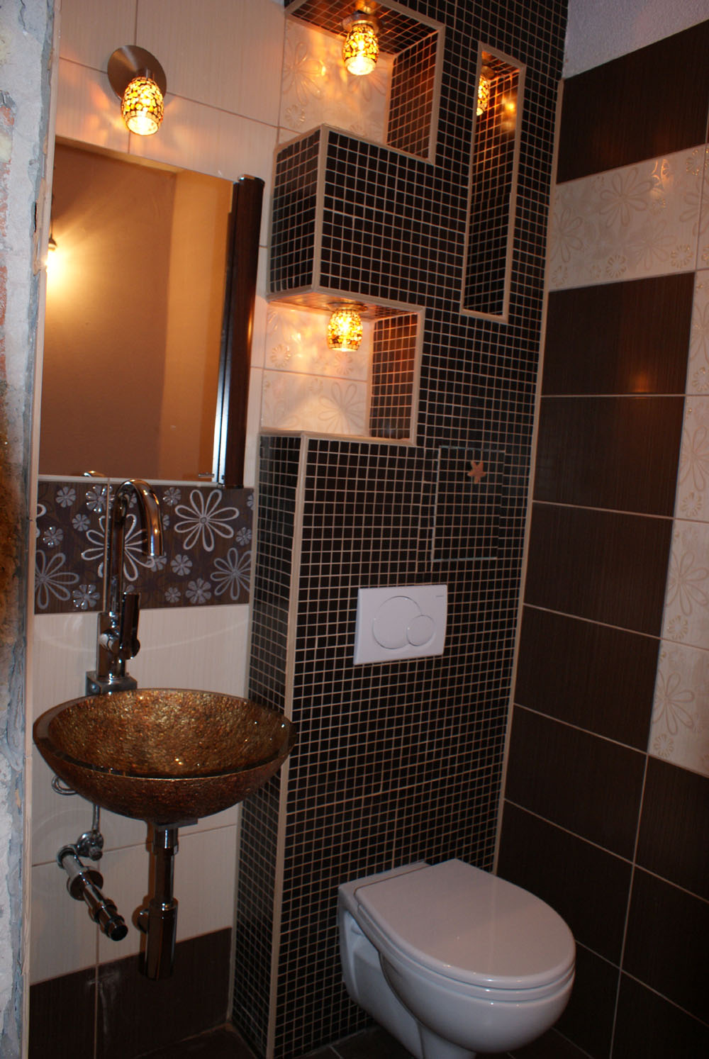 Umywalka w kolorze miodowym, zamontowana na postumencie, z chromowaną baterią i dekoracyjnym wzorem kwiatowym na ścianie, oświetlona ozdobnymi lampkami, z toaletą w tle i mozaiką z czarnych płytek.
