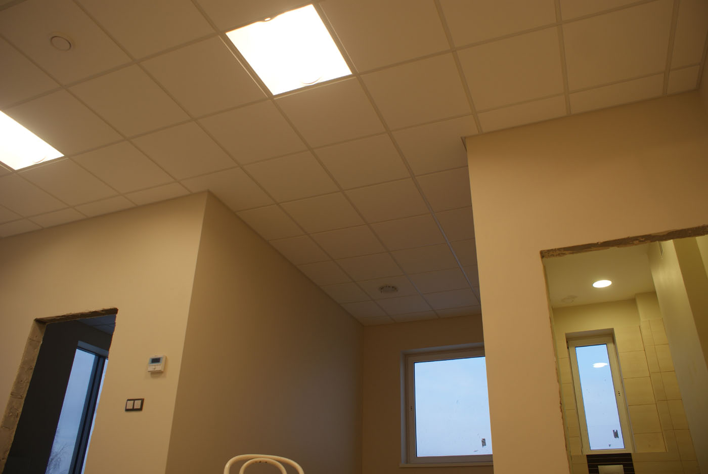 Sufit podwieszany kasetonowy z oświetleniem LED w nowo wykończonym pomieszczeniu biurowym, widoczne okna i otwór drzwiowy w trakcie wykańczania.