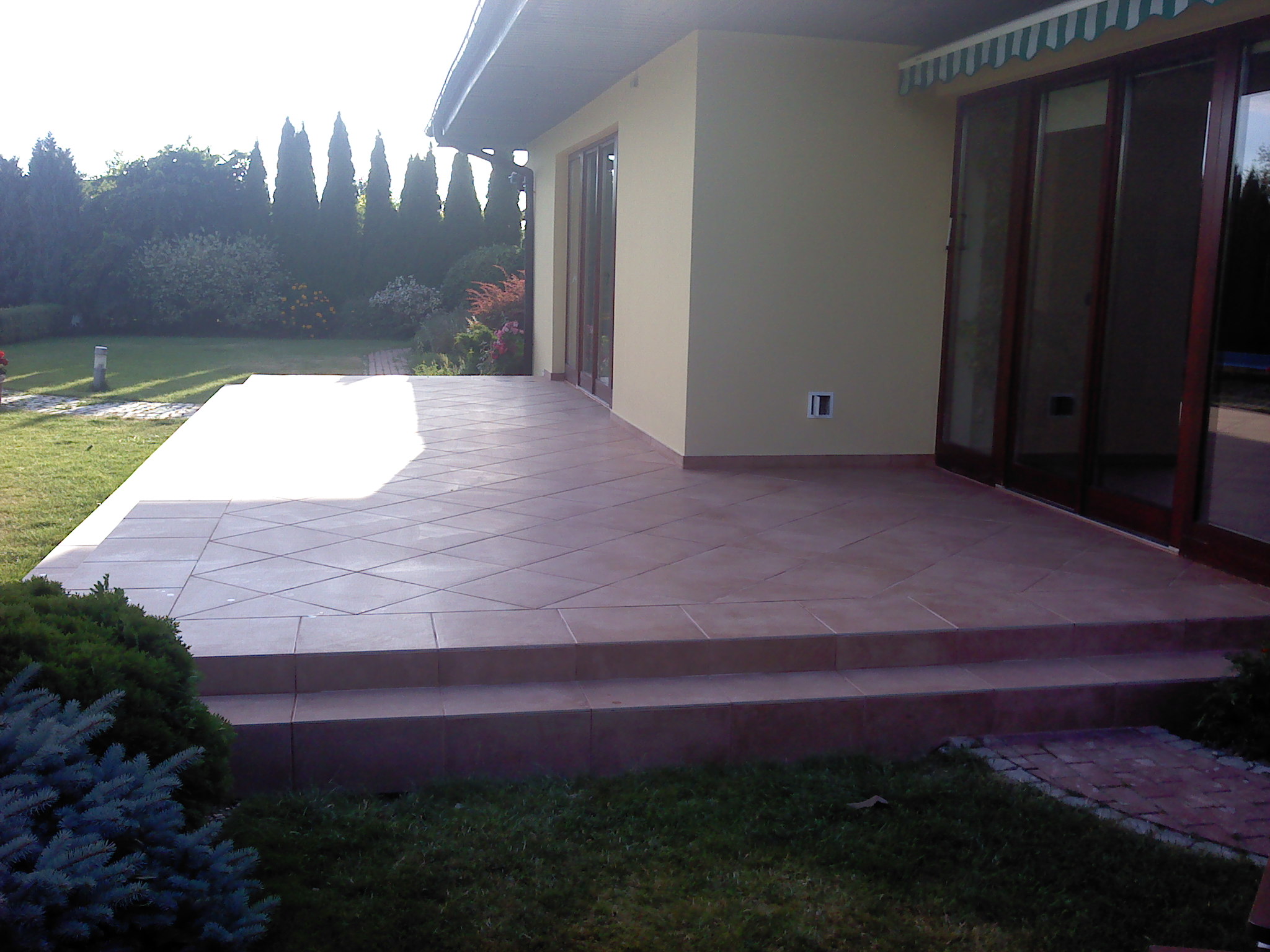 Wyłożony terakotą taras z dwoma stopniami, z widokiem na zadbany ogród z tujami i kwiatami, oświetlony popołudniowym słońcem.