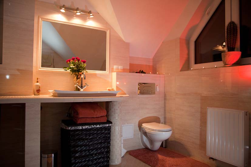 Jasna łazienka na poddaszu z beżowymi płytkami, umywalką z bukietem czerwonych róż, toaletą podtynkową i grzejnikiem panelowym.