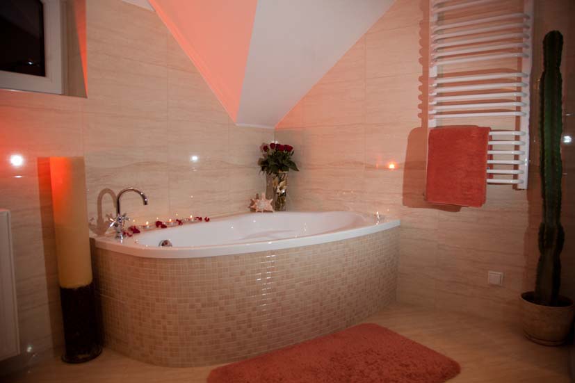 Jasna łazienka z narożną wanną obłożoną mozaiką, ozdobiona świecami i płatkami róż, grzejnik drabinkowy z pomarańczowym ręcznikiem, wysoki kaktus w doniczce.