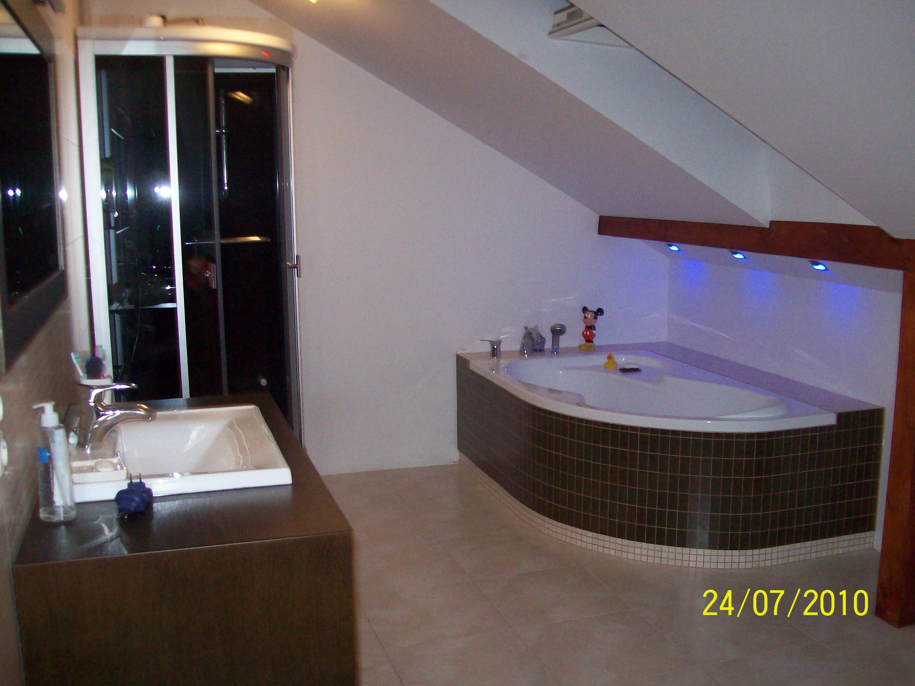 Wykończona łazienka na poddaszu z kabiną prysznicową, umywalką z brązowym blatem i wanną z hydromasażem wypełnioną wodą, w której pływa gumowa kaczuszka i figurka Myszki Miki.