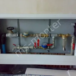 Wnęka z rurami, zaworami, wodomierzami i filtrem, zamknięta białą ramą w ścianie, widoczne podłączenia hydrauliczne.