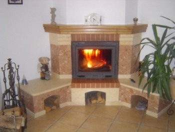 Kominek obudowany marmurkową płytką z ceglastymi akcentami, z drewnianą półką nad paleniskiem, na którym stoi zegar. W palenisku płonące drewno. Obok kominka dekoracje i roślina doniczkowa.