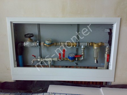 Wnęka z rurami, zaworami, wodomierzami i filtrem, zamknięta białą ramą w ścianie, widoczne podłączenia hydrauliczne.