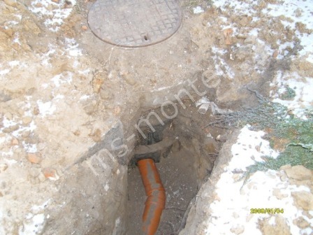 Wykop z widocznym fragmentem rury kanalizacyjnej z tworzywa sztucznego, wystającej spod ziemi, przykryty włazem kanalizacyjnym, w zimowej scenerii.