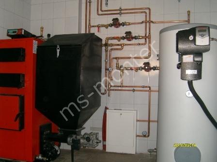 Kotłownia z kotłem na paliwo stałe z podajnikiem, zbiornikiem na wodę i instalacją z rur miedzianych na tle białych płytek.