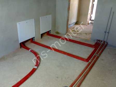 Czerwone rury centralnego ogrzewania ułożone na surowym podłożu w nowym budynku, z białymi panelami rewizyjnymi na ścianie i widocznymi pionami instalacji.