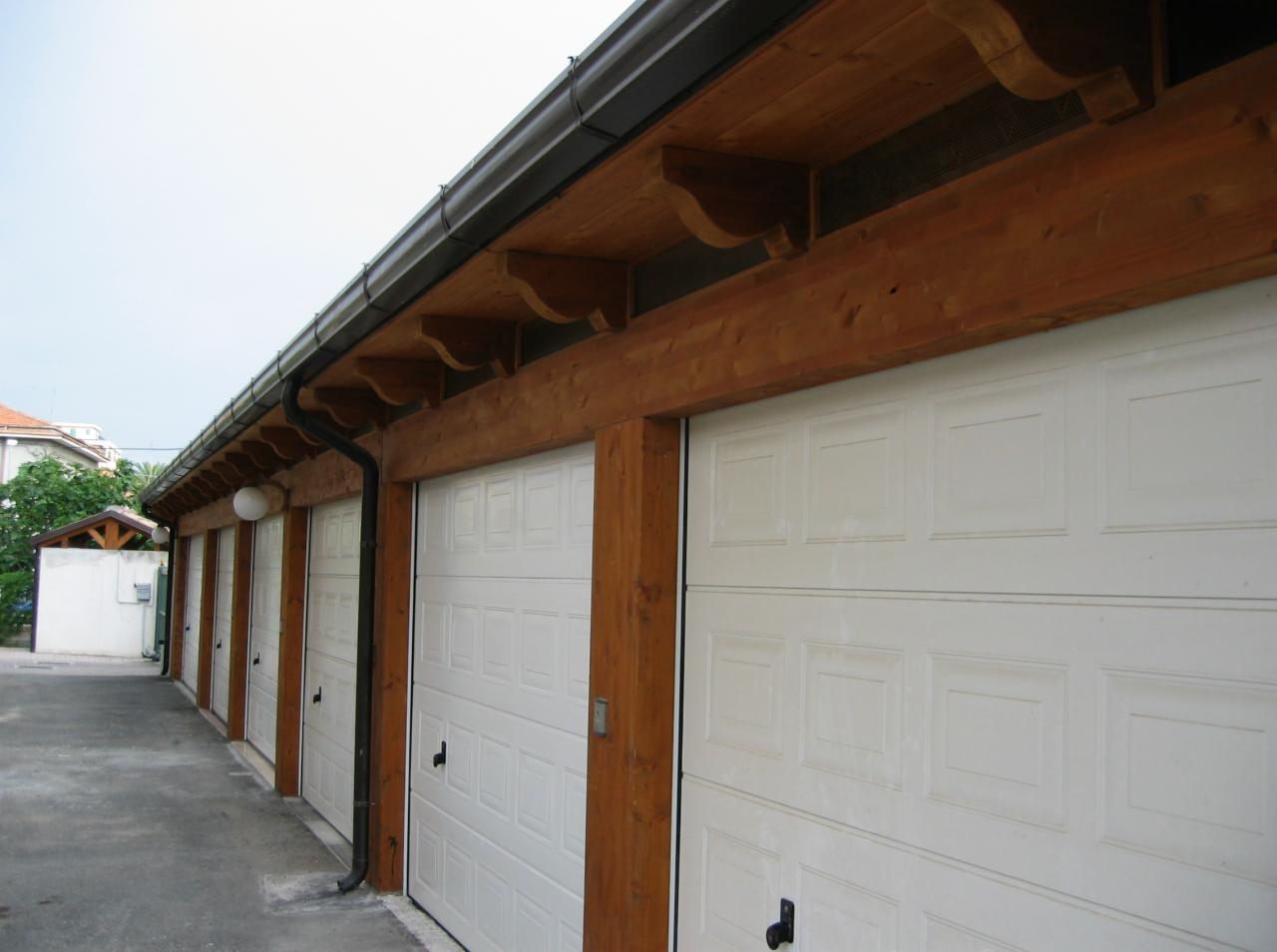 Rząd białych bram garażowych z brązową drewnianą konstrukcją zadaszenia i ozdobnymi wspornikami, z czarną rynną i rurą spustową po lewej stronie.