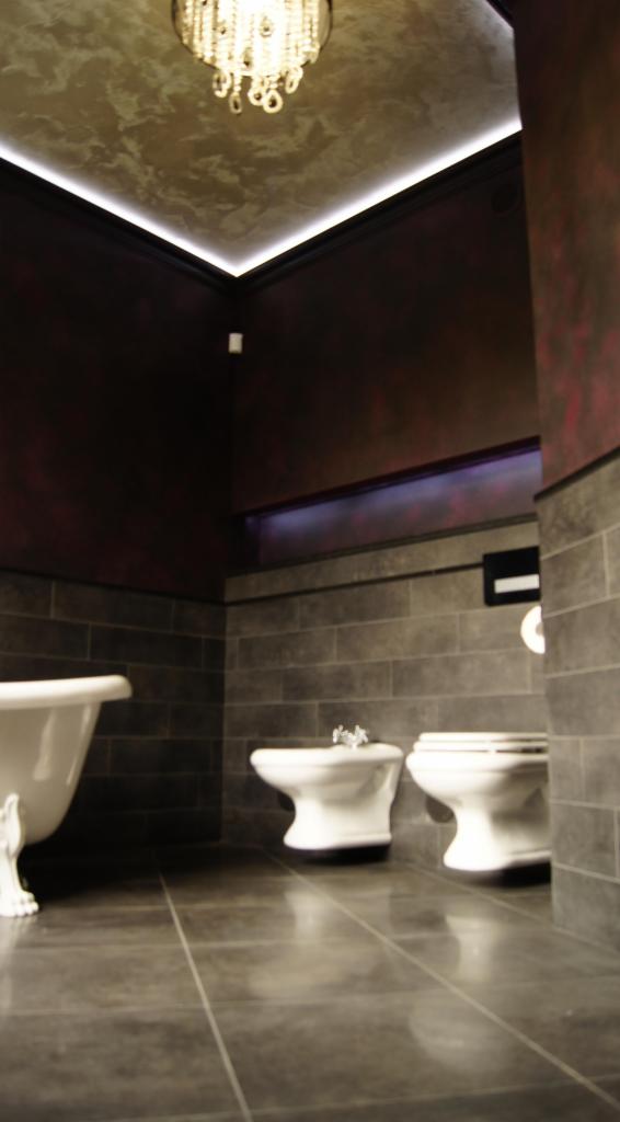 Elegancka łazienka w ciemnych tonacjach z wanną na nóżkach, bidetem i toaletą, oświetlona ozdobnym żyrandolem i listwą LED pod sufitem, ściany wykończone płytkami imitującymi kamień i dekoracyjnym...