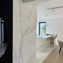 Nowoczesne wnętrze z marmurowymi płytami na ścianach i podłodze, widoczna kuchnia z wyspą i złotymi hokerami, czarny element dekoracyjny z lustrem, drewniana podłoga w części jadalnej.