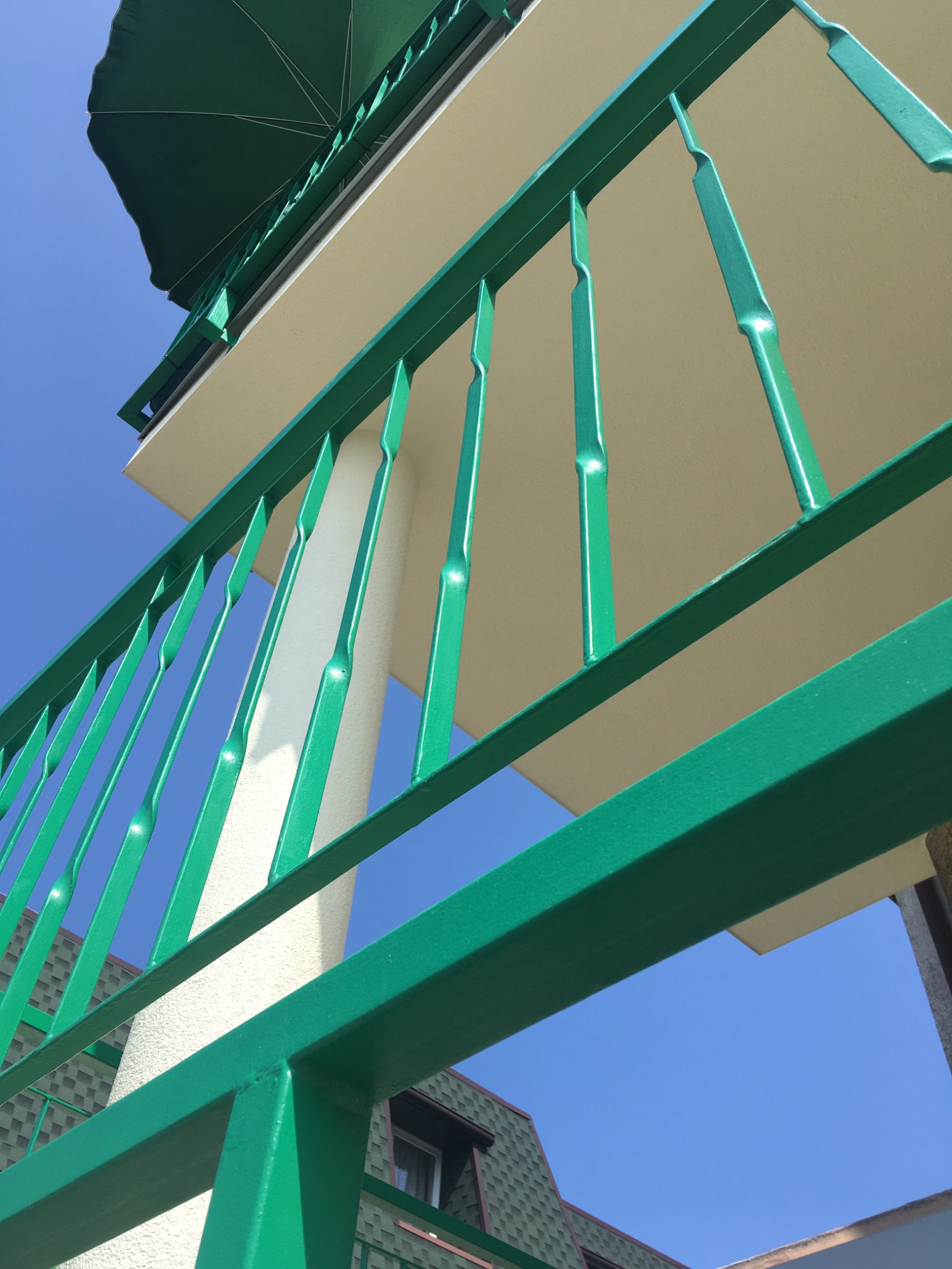 Zielona, metalowa balustrada balkonowa z nietypowymi, wygiętymi pionowymi elementami, widziana z dołu na tle błękitnego nieba i fragmentu budynku z zielonym dachem.