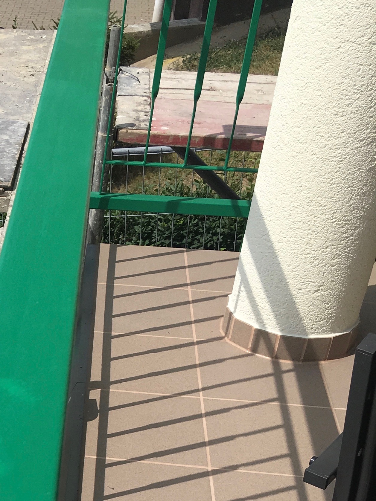 Fragment balkonu z zieloną balustradą, kolumną o chropowatej fakturze i cieniowanymi płytkami podłogowymi w ciepłym odcieniu.