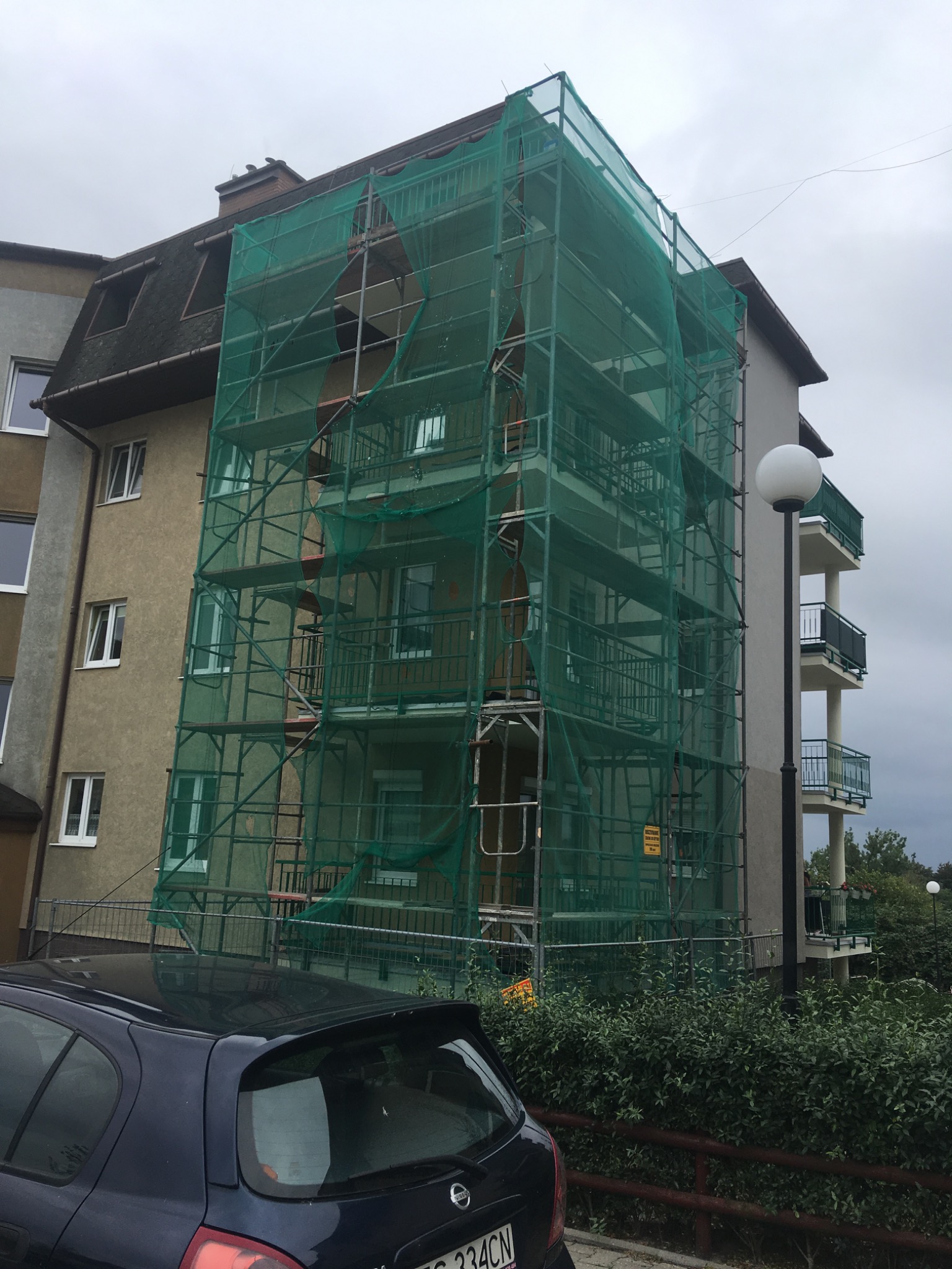 Wielopiętrowy budynek mieszkalny w trakcie prac remontowych, osłonięty rusztowaniem z zieloną siatką zabezpieczającą, widoczny fragment zaparkowanego samochodu i oświetlenie uliczne.