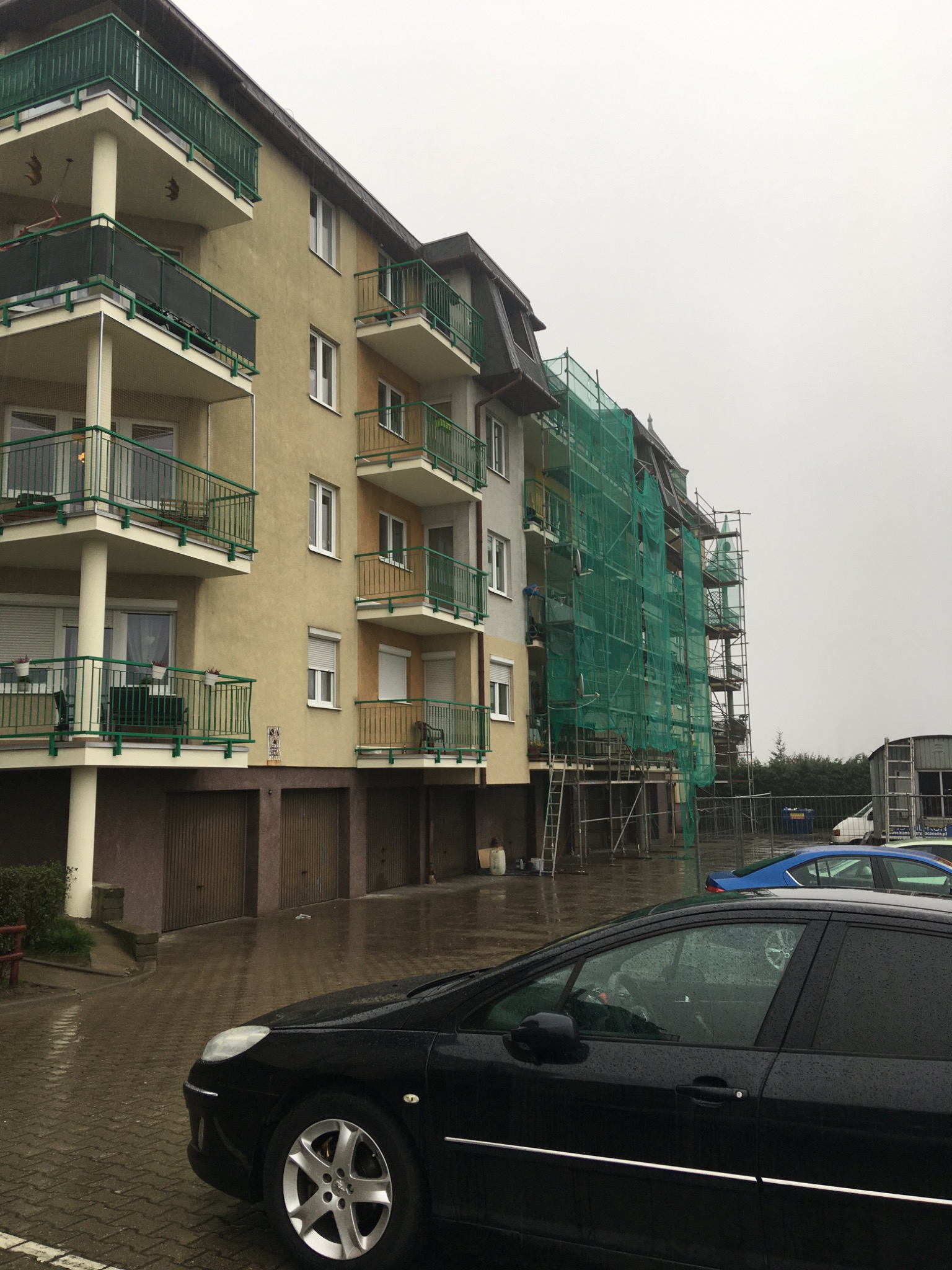 Wielopiętrowy budynek z balkonami, częściowo zasłonięty rusztowaniami i zieloną siatką zabezpieczającą podczas prac remontowych, widok z poziomu mokrego parkingu z zaparkowanym czarnym samochodem.