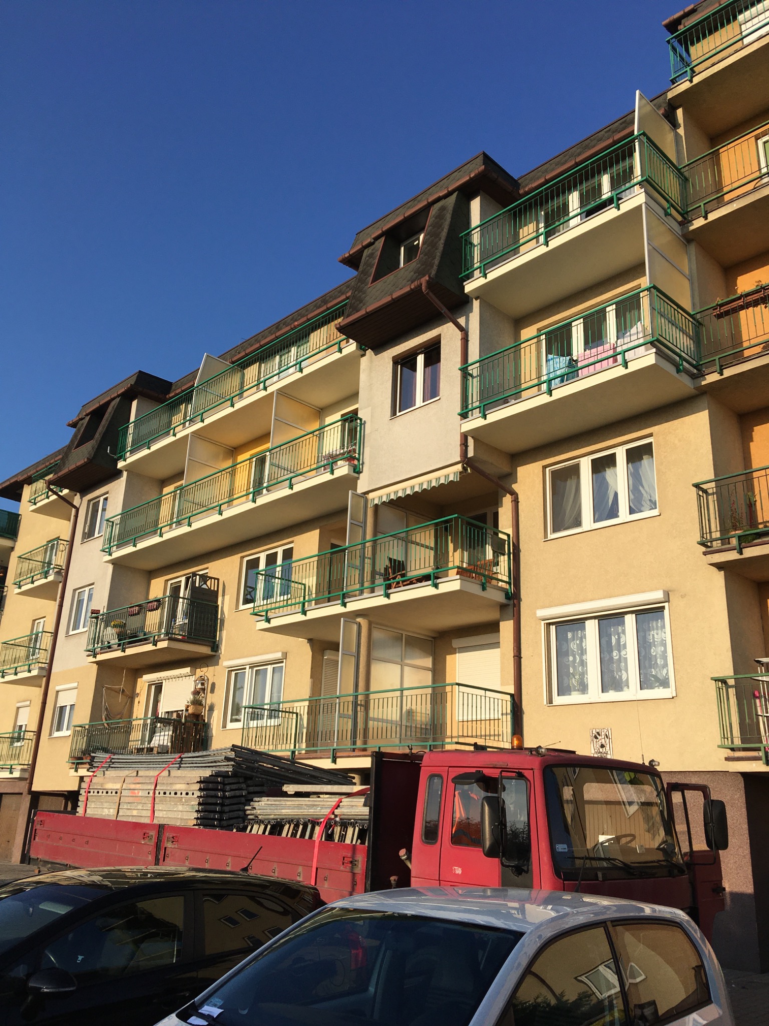 Wielopiętrowy budynek z balkonami i zielonymi balustradami, na pierwszym planie czerwona ciężarówka z załadunkiem rusztowań.