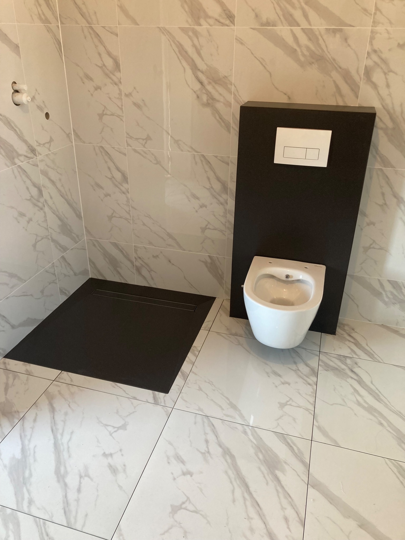 Nowoczesna łazienka z podwieszanym WC z czarną obudową i odpływem liniowym w kabinie prysznicowej, wykończona białymi płytkami imitującymi marmur.