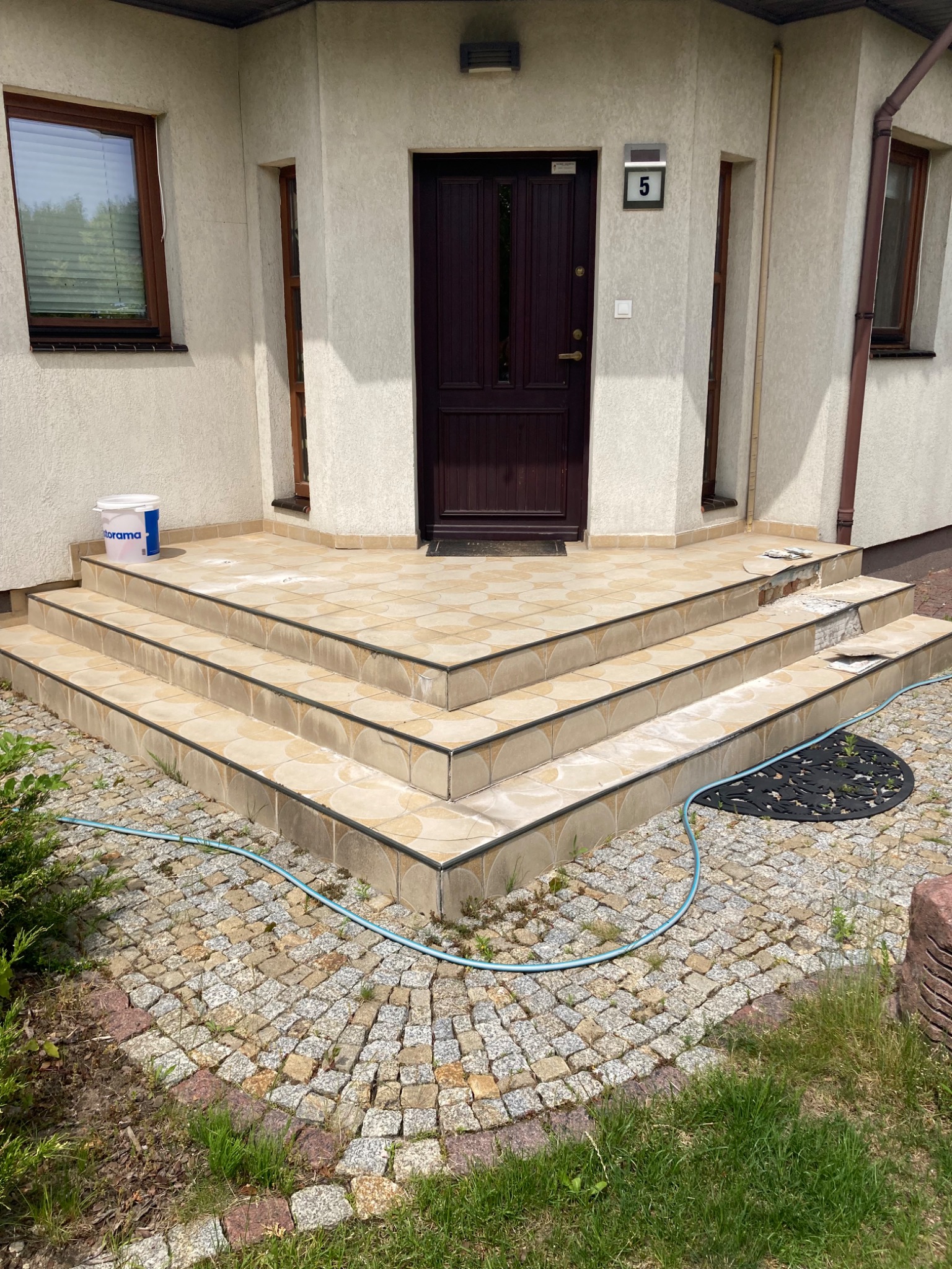 Taras przed domem z uszkodzonymi płytkami na schodach, widoczny częściowy demontaż płytek, obok stoi wiadro z farbą. Brukowane podejście.