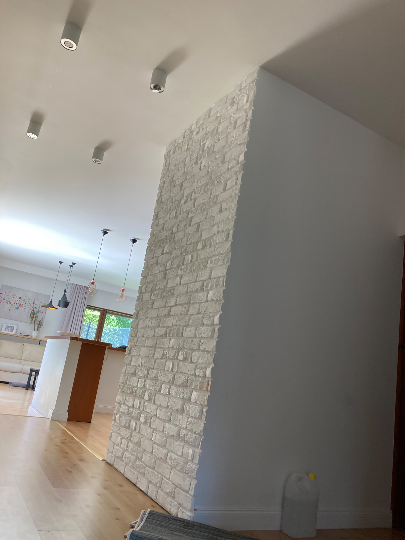 Wnętrze domu z fragmentem ściany wykończonej białą cegłą dekoracyjną, widoczne oświetlenie sufitowe, podłoga z jasnego drewna oraz fragment kuchni w tle.