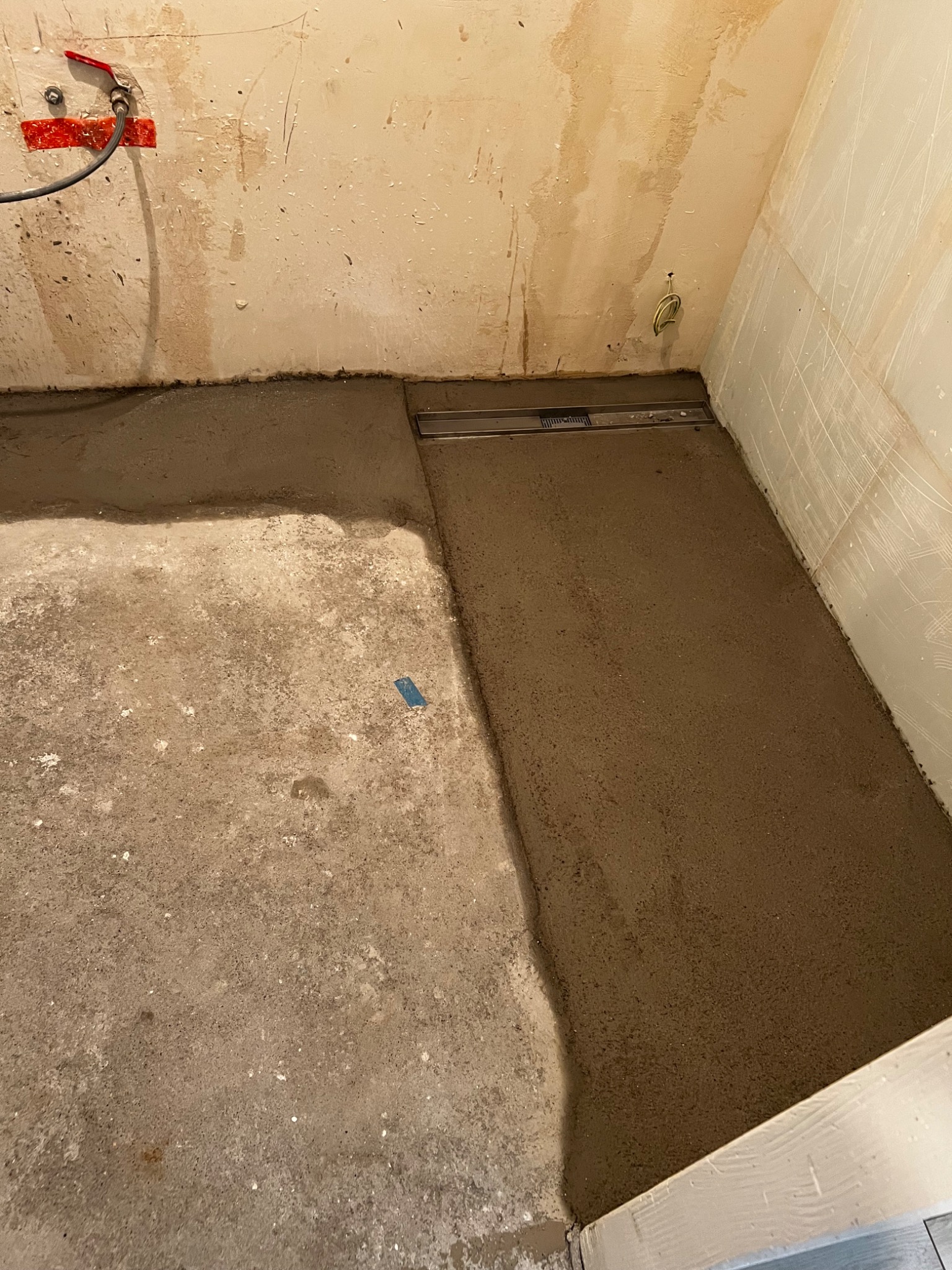 Przygotowanie podłoża pod prysznic: wylewka betonowa z odpływem liniowym na nierównym podłożu, widoczne ślady po instalacji hydraulicznej na ścianie.