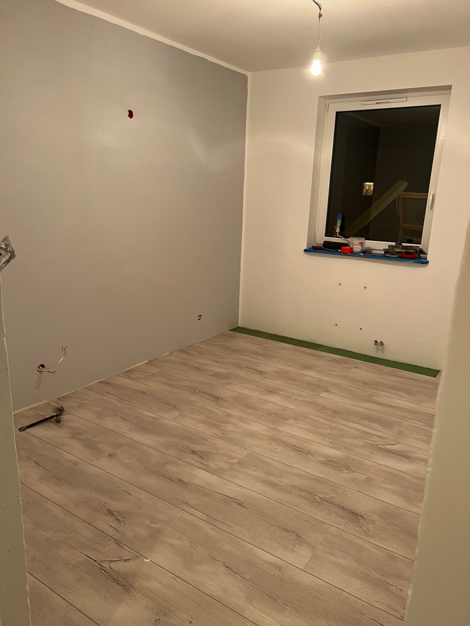 Pokój w trakcie remontu: świeżo położona podłoga laminowana, ściany częściowo pomalowane na szaro, widoczne puszki elektryczne i tymczasowe oświetlenie żarówką.