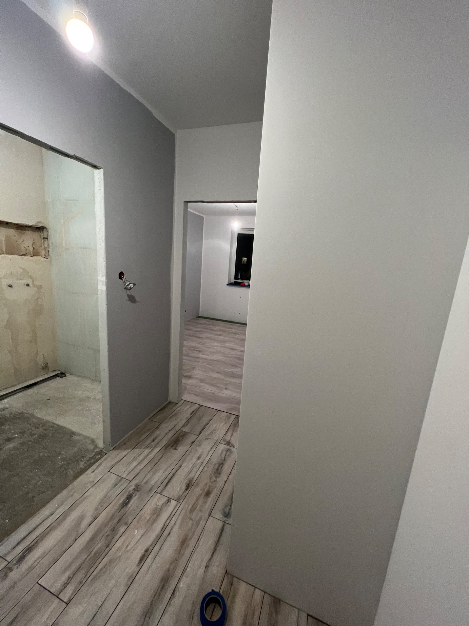 Wnętrze w trakcie remontu: świeżo położone płytki podłogowe imitujące drewno, szare ściany częściowo pomalowane, widoczne fragmenty instalacji elektrycznej i stan surowy w sąsiednim pomieszczeniu.